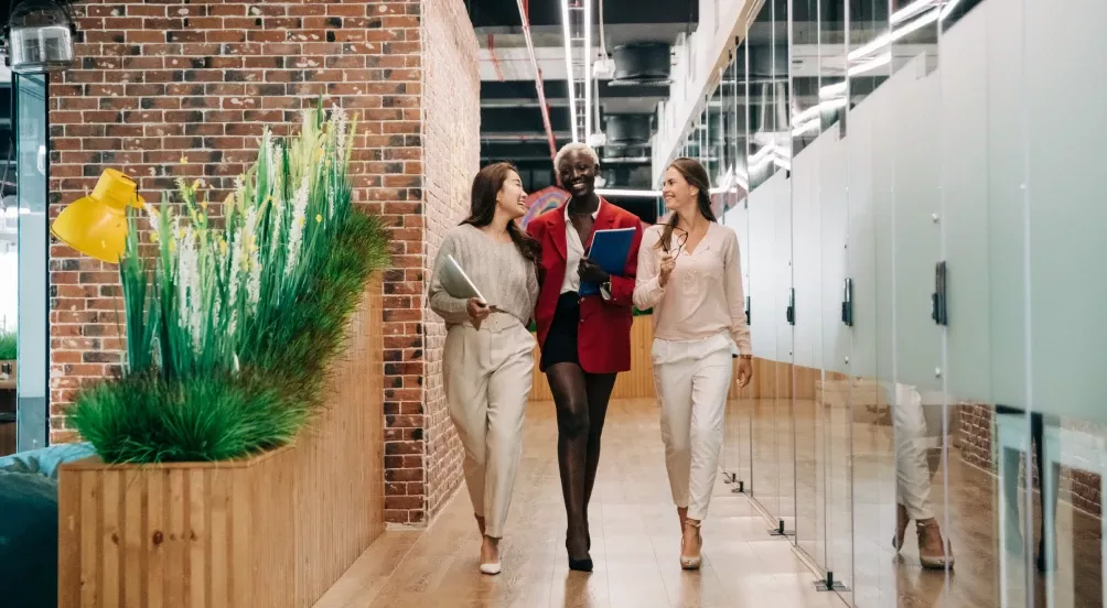 Tre personer går sammen gjennom en moderne kontorkorridor, prater og smiler, omgitt av glassvegger, grønne planter og teglvegger.